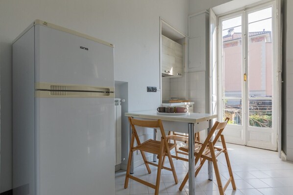 A compact kitchen with a white refrigerator, a dining table with chairs, and a view through an open door to the outside.