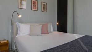 Ein ordentlich bezogenes Bett mit weißen und rosafarbenen Kissen, einer dunklen Decke und einer Nachttischlampe. Darüber an der Wand hängen drei gerahmte Bilder.