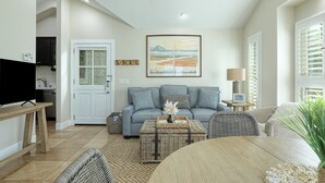 En moderne stue med sofa, sofabord og fjernsyn. Et maleri hænger på væggen, og et vindue har persienner.