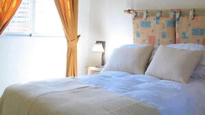 Un lit recouvert d’une courtepointe blanche, une table de chevet avec une lampe et une fenêtre avec des rideaux.