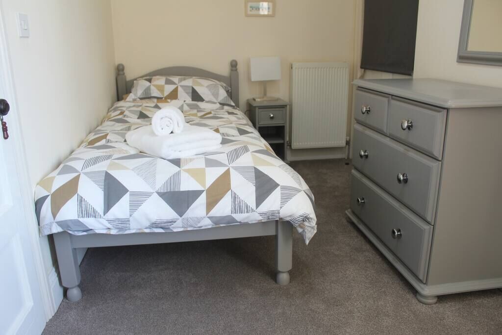 A neatly made bed with a geometric patterned bedspread and a rolled white towel. There is a bedside table with a lamp, a dresser with drawers, and a radiator on the wall.