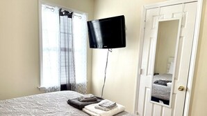 A bedroom with a bed, a flat-screen TV mounted on the wall, a door with a mirror, and a window with curtains. - Quiet & Downtown Smart TVs in all bedrooms, minutes from base. (Havelock)