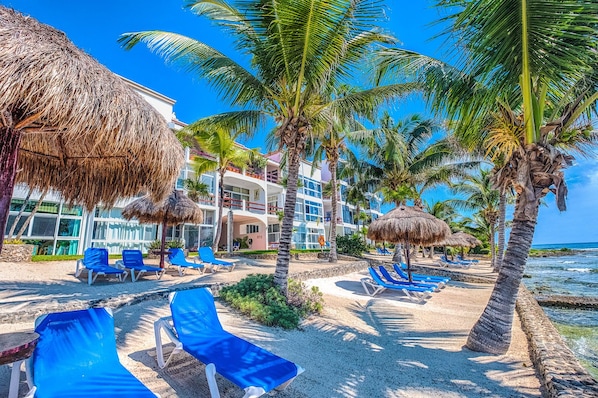 A beach resort with thatched umbrellas, palm trees, and blue lounge chairs.