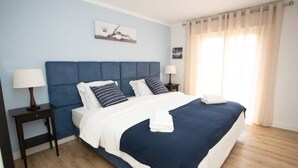 A neatly made bed with a blue headboard, white bedding, and striped pillows. There are two bedside tables with lamps, and a window with curtains. - Meriteluna Pateo - beetwen Lisboa Sintra e Cascais (Agualva-Cacém)