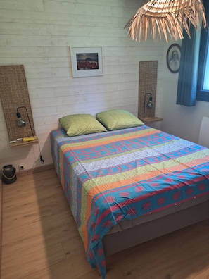A bedroom with a wooden floor, a bed with a colorful quilt, a woven wall hanging, and a wall-mounted lamp.