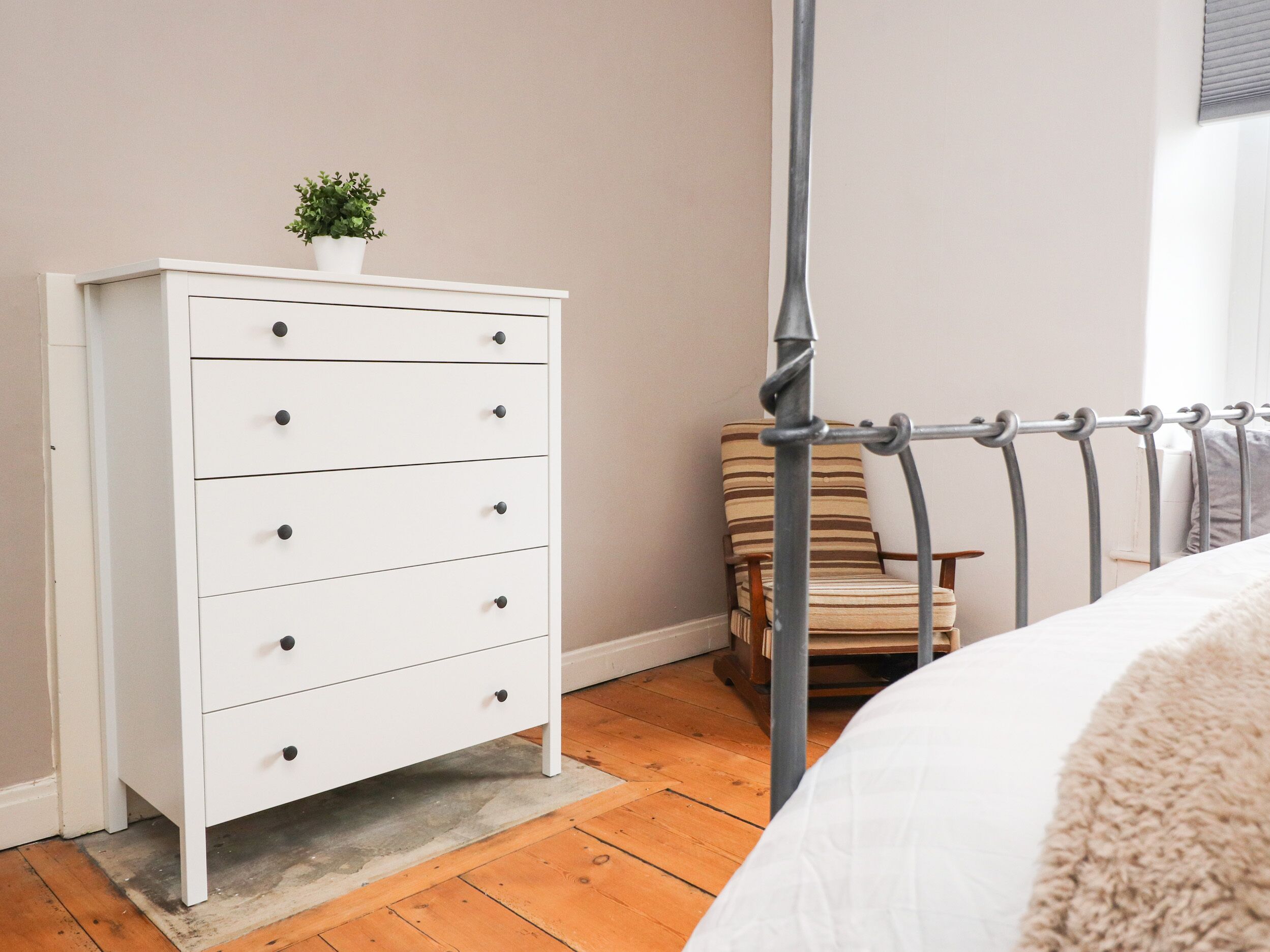 Un dormitorio con un tocador blanco, una silla y una estructura de cama de metal.