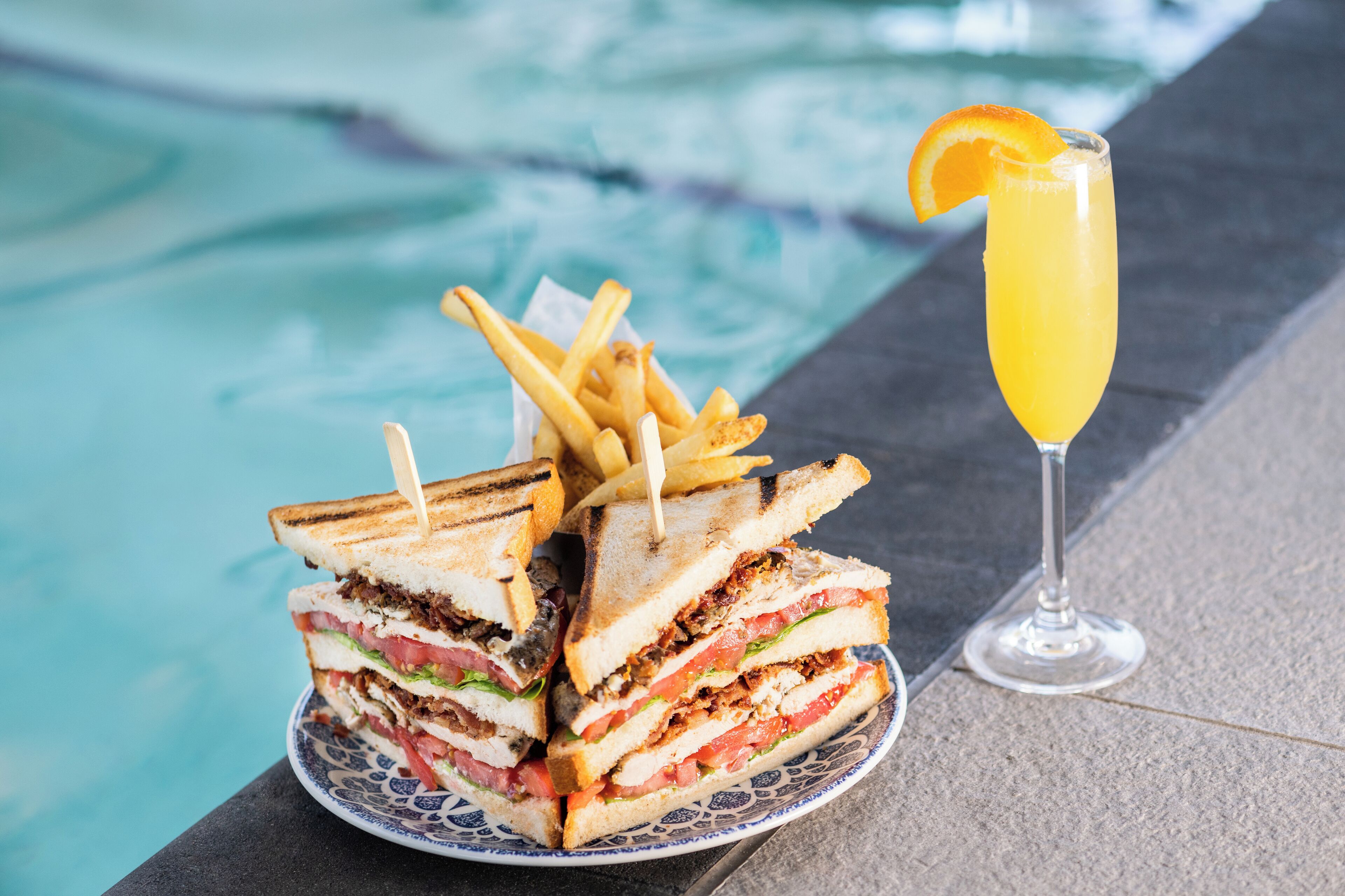 Une assiette de sandwichs grillés et de frites, à côté d’un verre de jus d’orange, près d’une piscine.