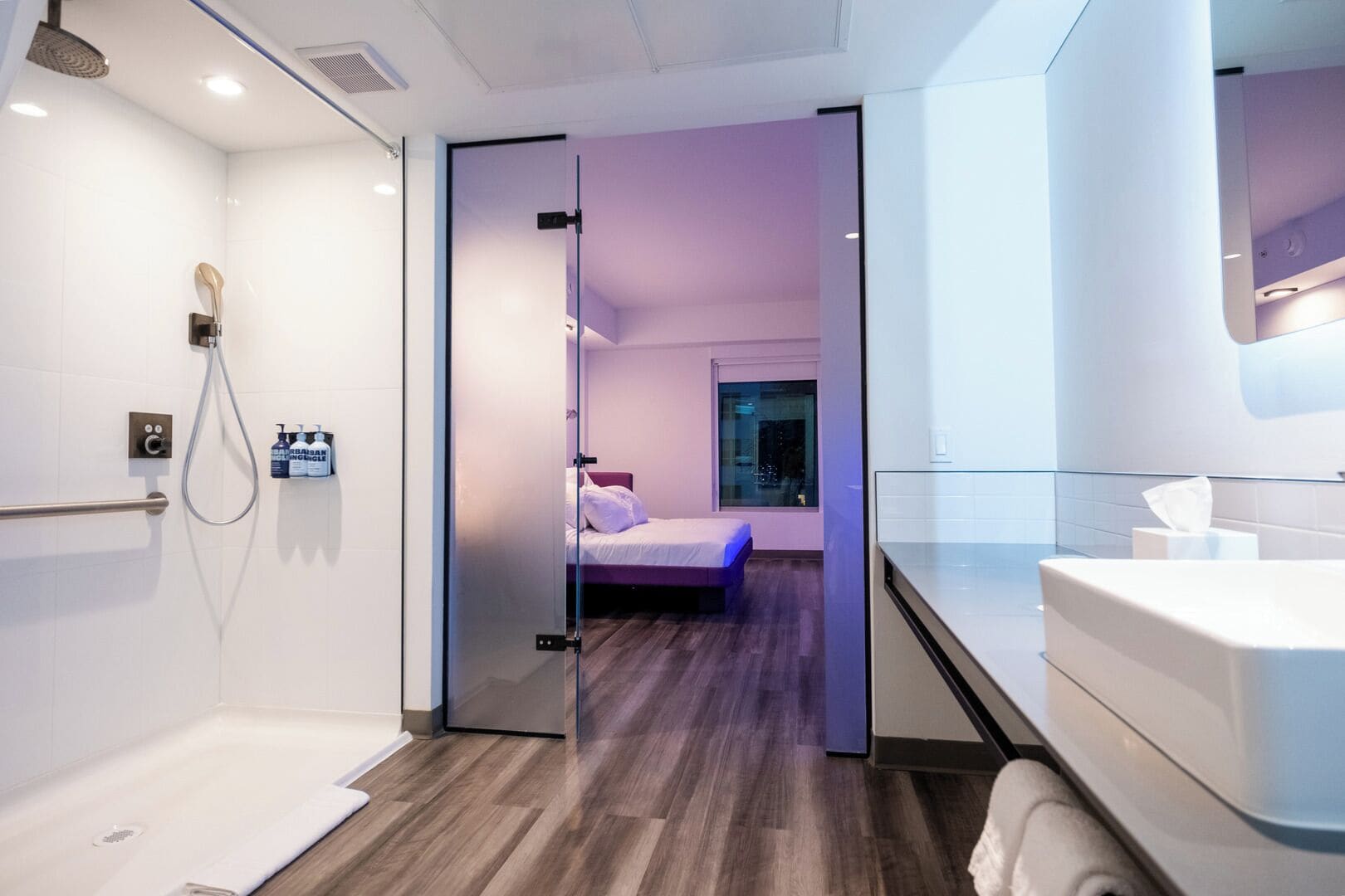 A modern bathroom with a shower, a toilet, and a sink. There is a bed visible through a glass door in the adjacent room.