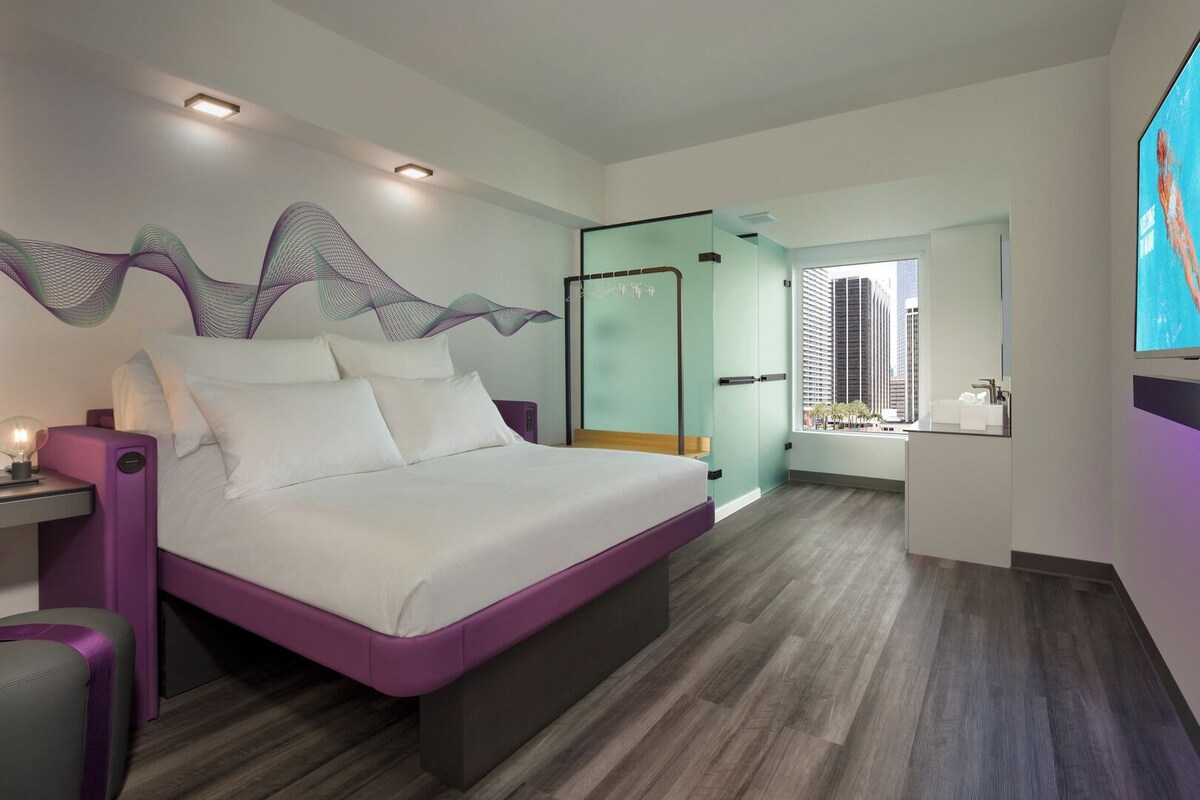 A modern hotel room with a large bed, a bedside table, a TV, and a view of the city.