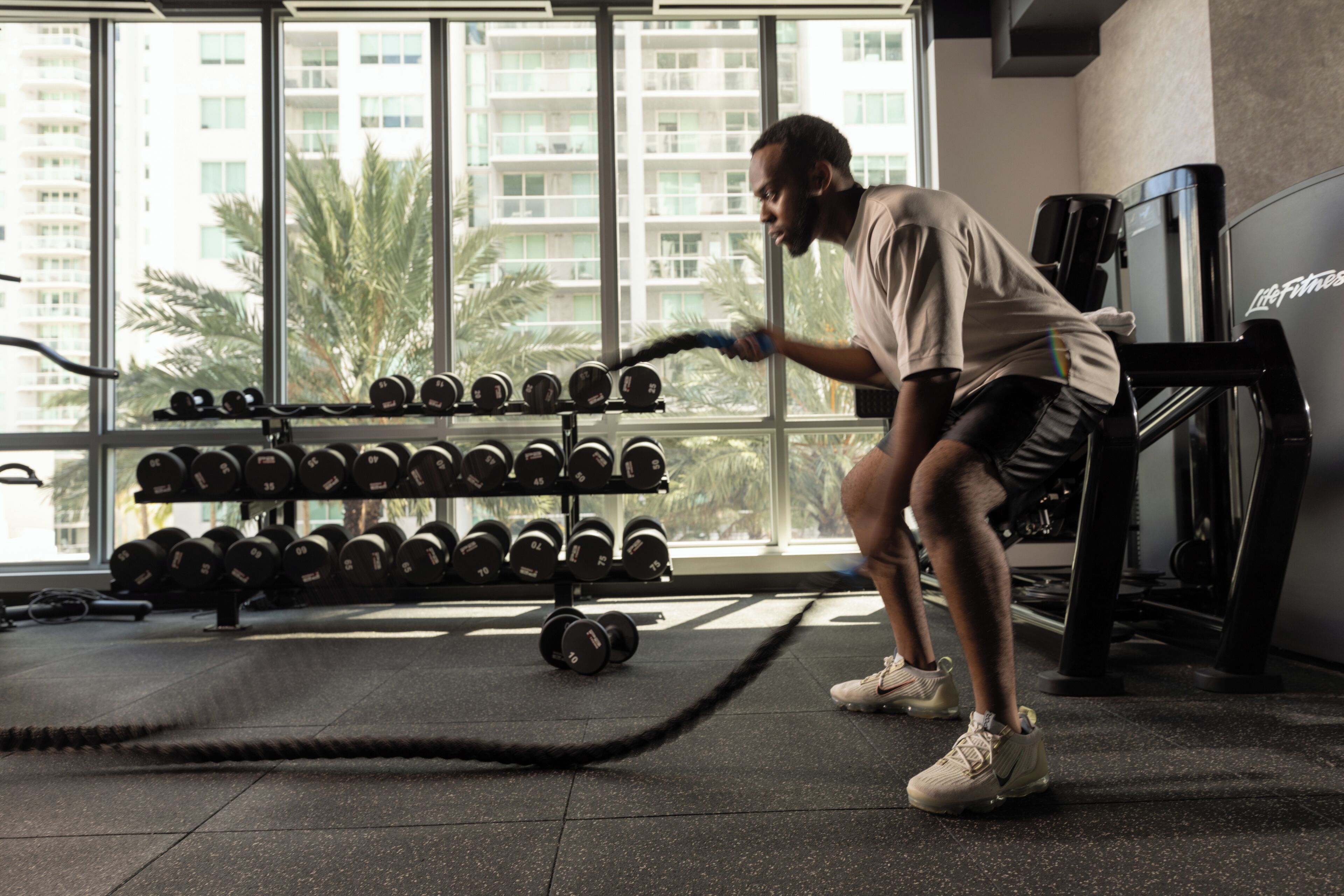 A gym with a person lifting weights, a rack of dumbbells, and a large window with a view of buildings and palm trees.