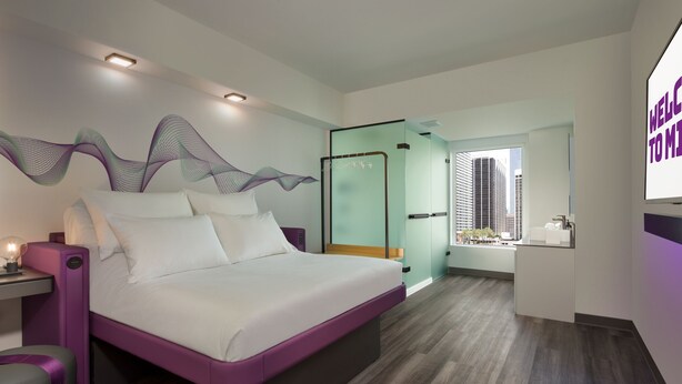 A modern hotel room with a large bed, a bedside table with a lamp, a glass partition, and a view of the city. - YOTEL Miami (Miami)