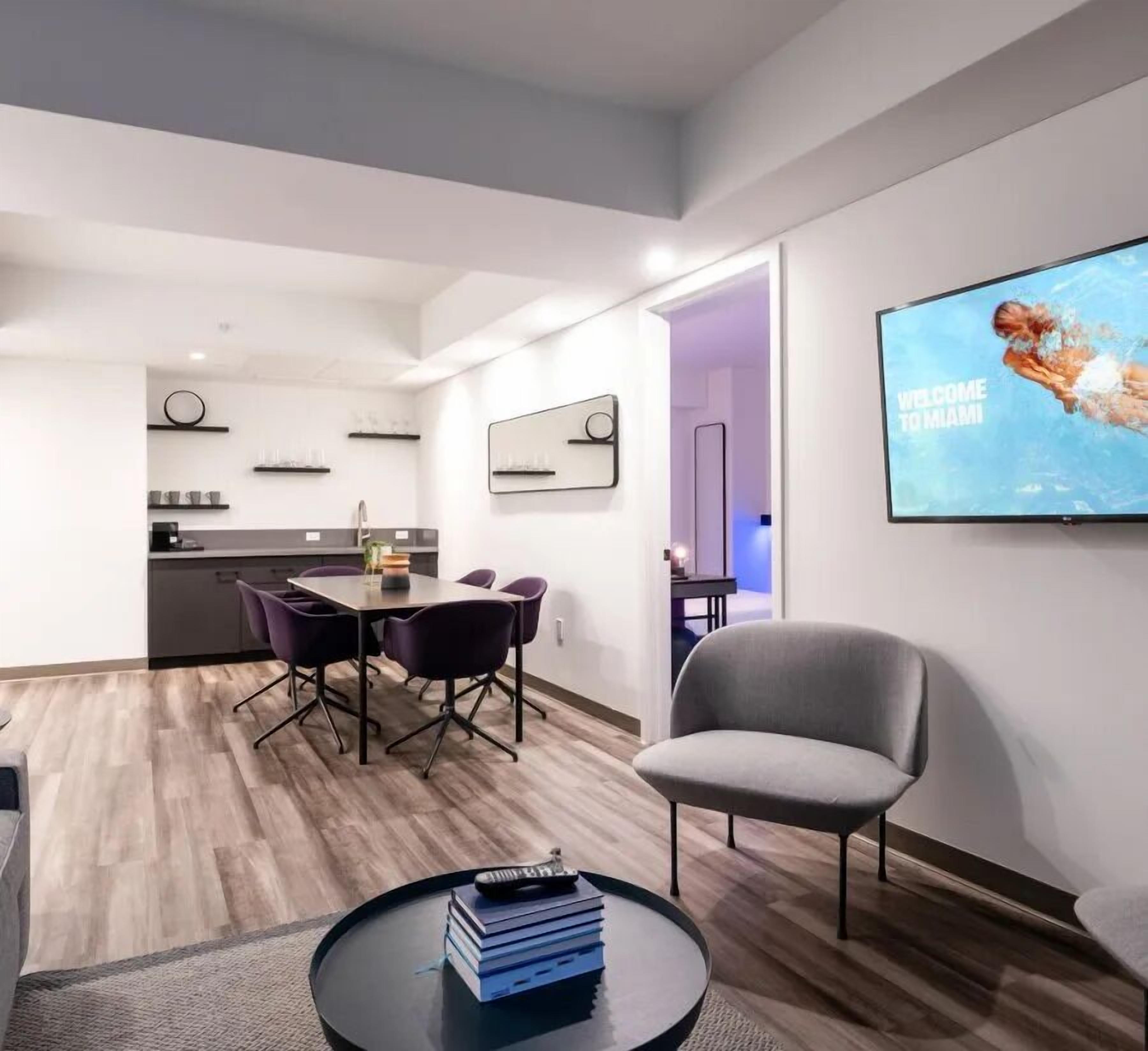 A modern living room with a flat-screen TV, a dining area with a table and chairs, and a kitchen in the background.