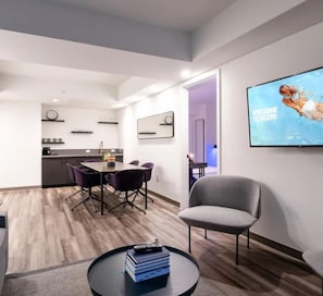 A modern living room with a flat-screen TV, a dining area with a table and chairs, and a kitchen in the background.
