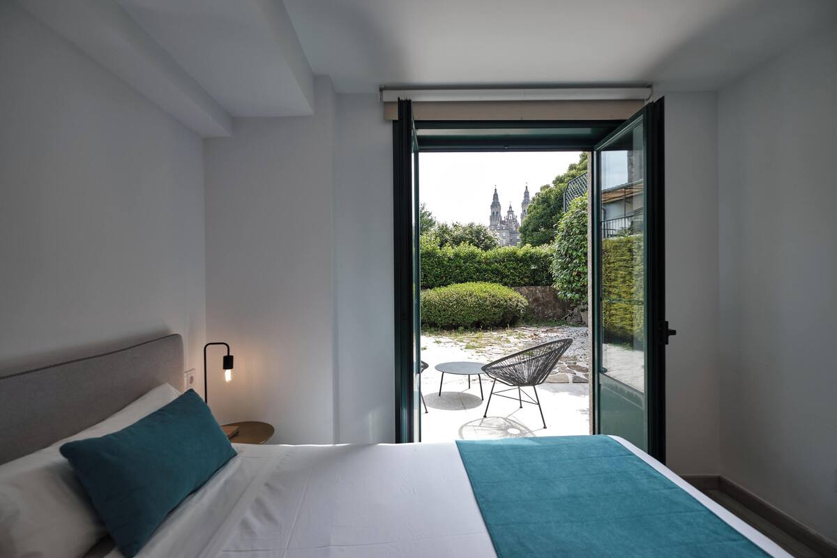 Ein modernes Hotelzimmer mit Bett, Nachttisch, Lampe und Blick auf Garten und Gebäude durch eine Glasdoor.