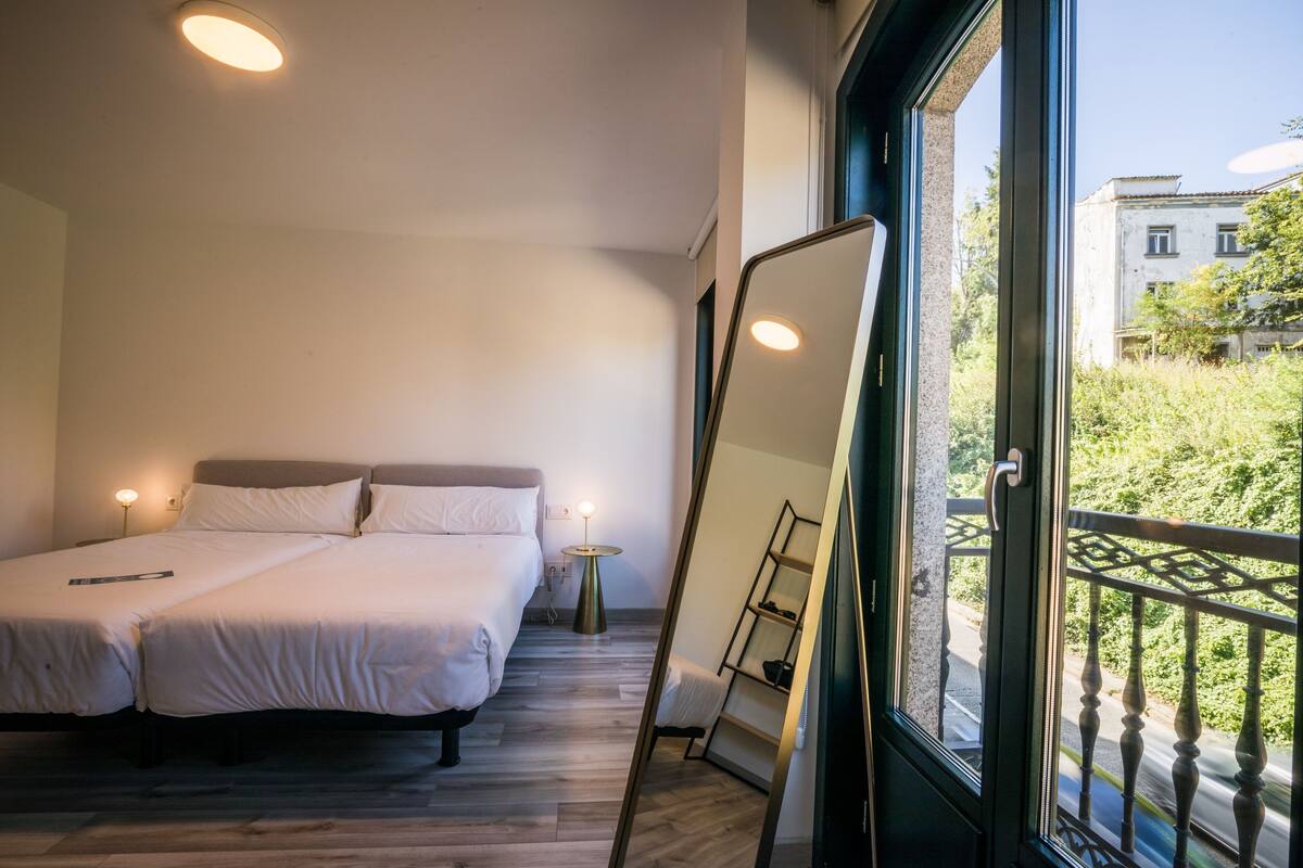Ein Schlafzimmer mit zwei Betten, einem Nachttisch mit Lampe, einem großen Spiegel und einem Balkon mit Geländer.
