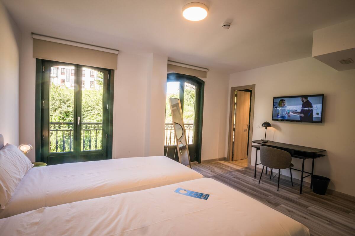 Ein Hotelzimmer mit zwei Betten, einem Schreibtisch, einem Fernseher und einem Balkon mit Blick auf Grünflächen.