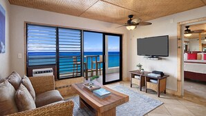 Una sala de estar con un gran ventanal corredera que da a un balcón con vistas al océano. Hay una mesa de centro de madera, un sofá y un televisor montado en la pared.