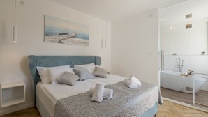 Ein modernes Schlafzimmer mit einem großen Bett, einem Gemälde an der Wand und einem durch eine Glasscheibe abgetrennten Badezimmer.