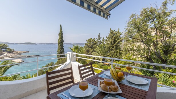 Ein Esstisch, gedeckt für das Frühstück, mit Croissants, Obst und Saftgläsern, mit Blick auf die Küste, Bäume und einen klaren Himmel.