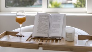 Ein Fenster mit Blick auf eine Küstenlandschaft, ein Buch, ein Glas bernsteinfarbene Flüssigkeit und eine weiße Kerze auf einem Holftablett.