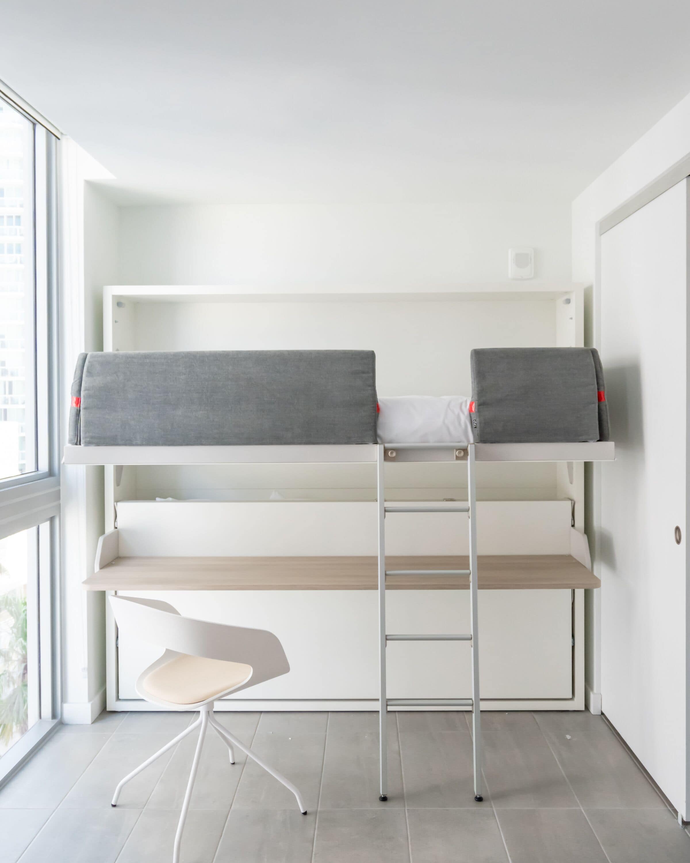 Una cama individual con ropa de cama gris, un escritorio pequeño y una silla en una habitación con un ventanal grande.