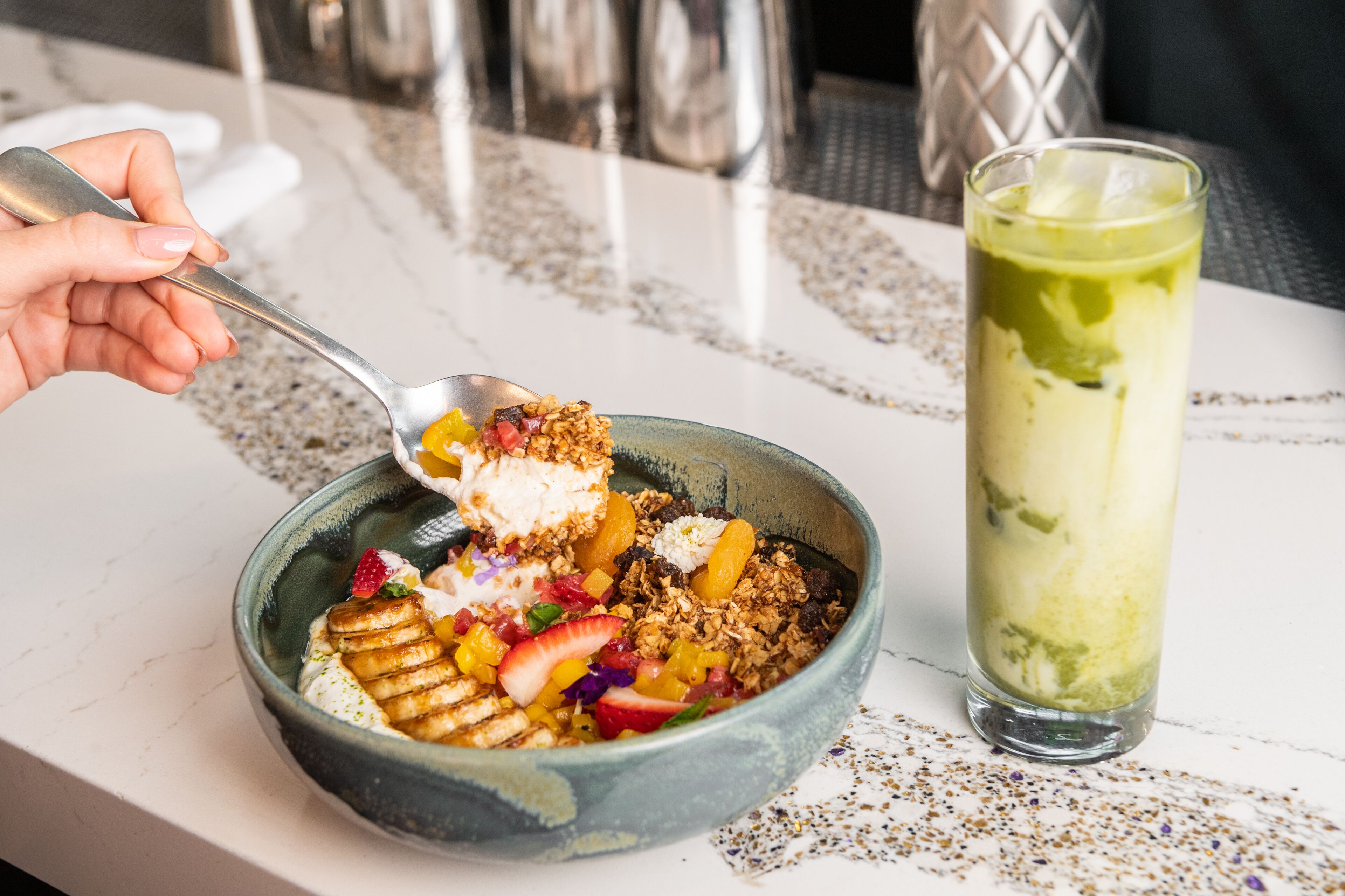 A bowl of food with a spoon, a glass of green drink, and a patterned tablecloth.
