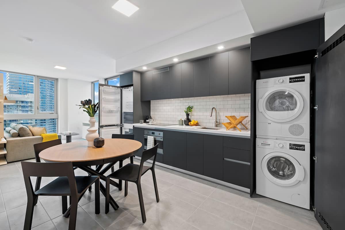 Une cuisine moderne avec des meubles de couleur sombre, un lave-linge et un s èche-linge intégrés, un coin repas avec une table ronde et des chaises, et une vue sur la ville grâce à de grandes fenêtres.