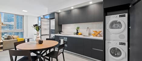 A modern kitchen with dark cabinetry, a built-in washer and dryer, a dining area with a round table and chairs, and a view of the city through large windows.