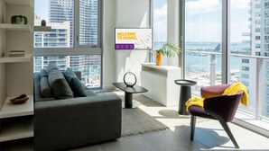 A modern living room with a grey sofa, a purple chair, a small table, and a TV displaying "WELCOME TO MIAMI."