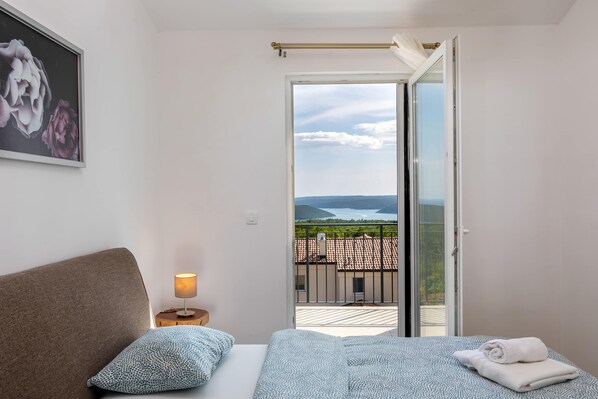 Ein Schlafzimmer mit Bett, Nachttisch, Lampe, Blick ins Freie und einer Tür zu einem Balkon.
