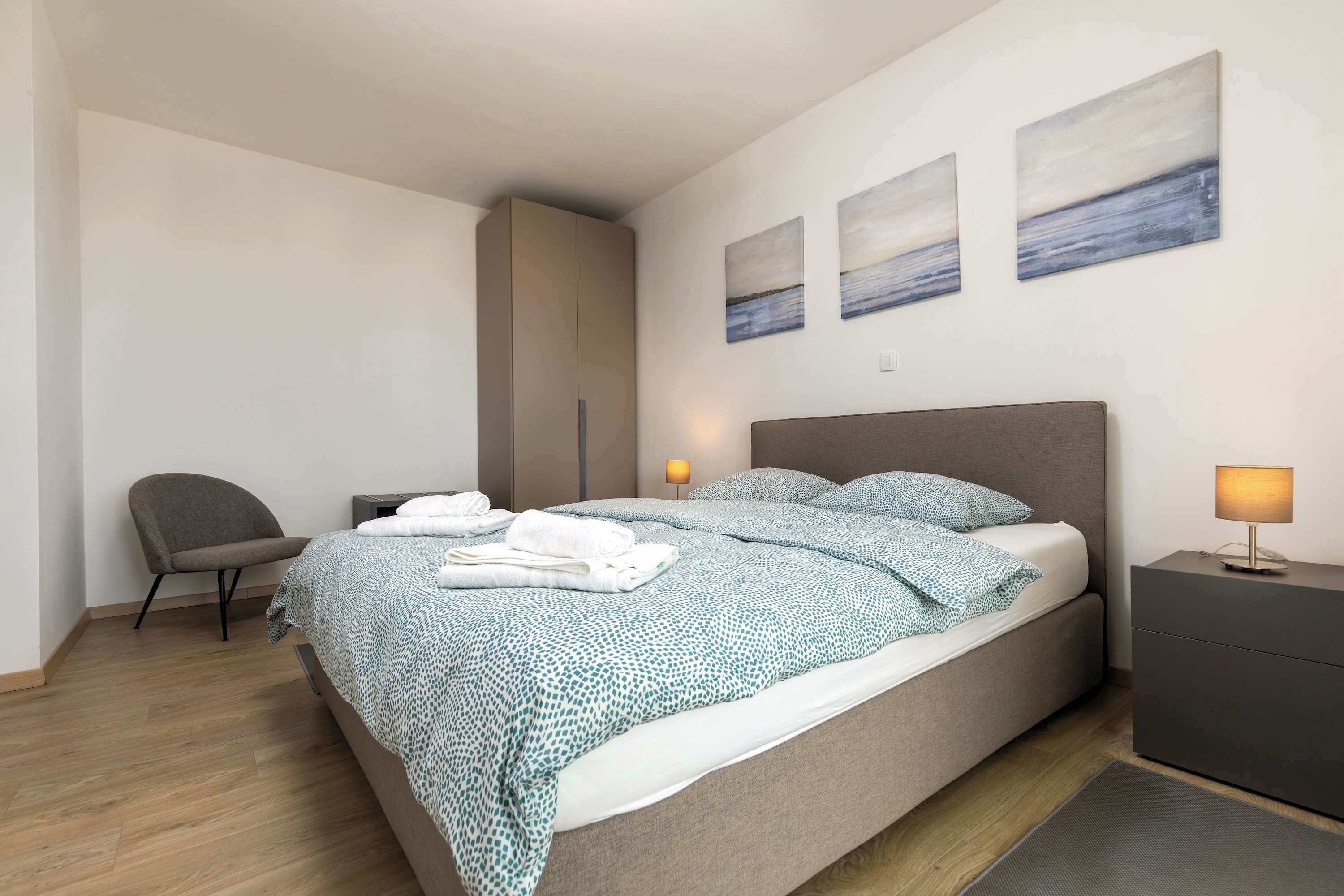 A modern bedroom with a bed, bedside tables, a chair, and artwork on the wall.