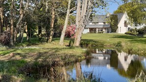 A house with a pond in front, surrounded by trees and a grassy lawn. - Elégant T2, 4⭐, cadre verdoyant, centre ville 750 m, plage et GR34 à 3 km. (Fouesnant)