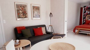 A modern living room with a black sofa, red and green cushions, a white coffee table, and a round woven tray. There are two framed floral paintings on the wall, a floor lamp, and a red bed in the background. - Elégant T2, 4⭐, cadre verdoyant, centre ville 750 m, plage et GR34 à 3 km. (Fouesnant)