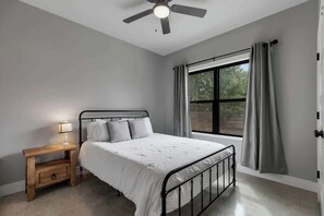A bedroom with a bed, a nightstand, a ceiling fan, a window with curtains, and a view of greenery outside.