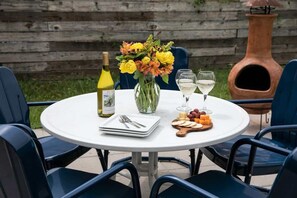 A round outdoor table with a white tablecloth, a bottle of white wine, a vase with yellow flowers, two wine glasses, a cheese board with crackers, and a set of cutlery.