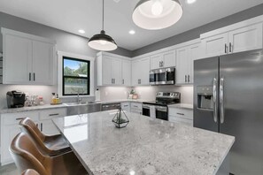 Une cuisine moderne avec des armoires blanches, des appareils en acier inoxydable, un grand îlot central et des luminaires suspendus.