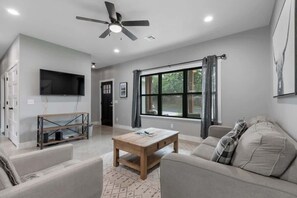 A modern living room with a flat-screen TV, a ceiling fan, a large window with curtains, and a beige sofa.