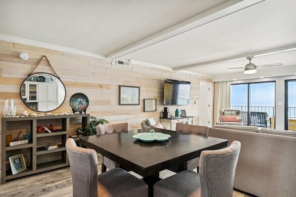 A modern living room with a wooden wall, a round mirror, a flat-screen TV, a dining table with chairs, and a balcony with a view.