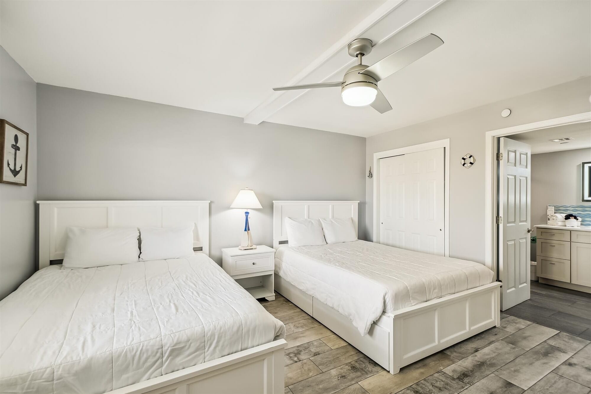 A bedroom with two beds, a ceiling fan, a nightstand with a lamp, and a bathroom visible through an open door.