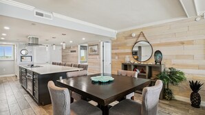 A modern kitchen with a central island, a dining area with a dark wooden table and grey upholstered chairs, and a wooden wall paneling. - Surf Dweller 610 is a Stunning Gulf Front 2 BR - sleep 6 by RedAwning (Fort Walton Beach)
