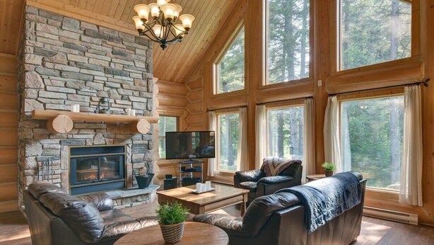 Ein gemütliches Blockhaus-Interieur mit Kamin aus Stein, Ledersofas, Fernseher und großen Fenstern mit Blick auf den Wald.