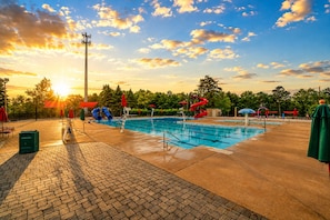 A swimming pool with slides and umbrellas, surrounded by trees and a sunset.