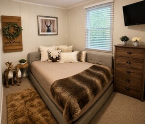 Une chambre à coucher comprenant un lit, une commode, un téléviseur, une fenêtre avec des stores, une couronne décorative accrochée au mur et un tableau représentant une tête de cerf encadrée.