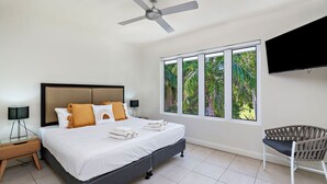 Ein Schlafzimmer mit einem Bett, einem Deckenventilator, einem Fernseher, einem Fenster mit Blick ins Grüne und einem Sessel.