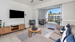 Ein modernes Wohnzimmer mit Flachbildfernseher, einer Holzunterhaltungsecke und einem Sofa mit Kissen. Es gibt einen Deckenventilator, einen kleinen runden Tisch und einen Sessel. Das Zimmer verfügt über eine große Schiebeglastür, die auf einen Balkon mit Blick auf Palmen und Gebäude führt.
