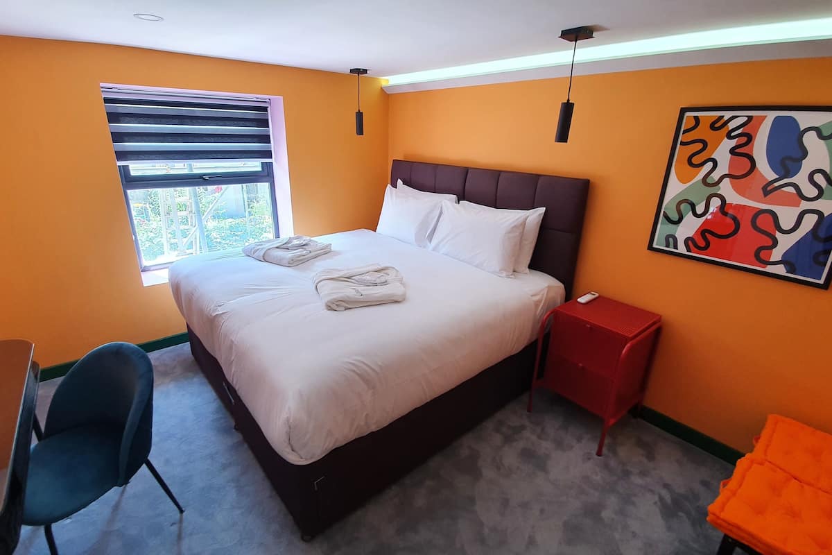 A hotel room with a bed, a desk, a chair, a nightstand, and a window.