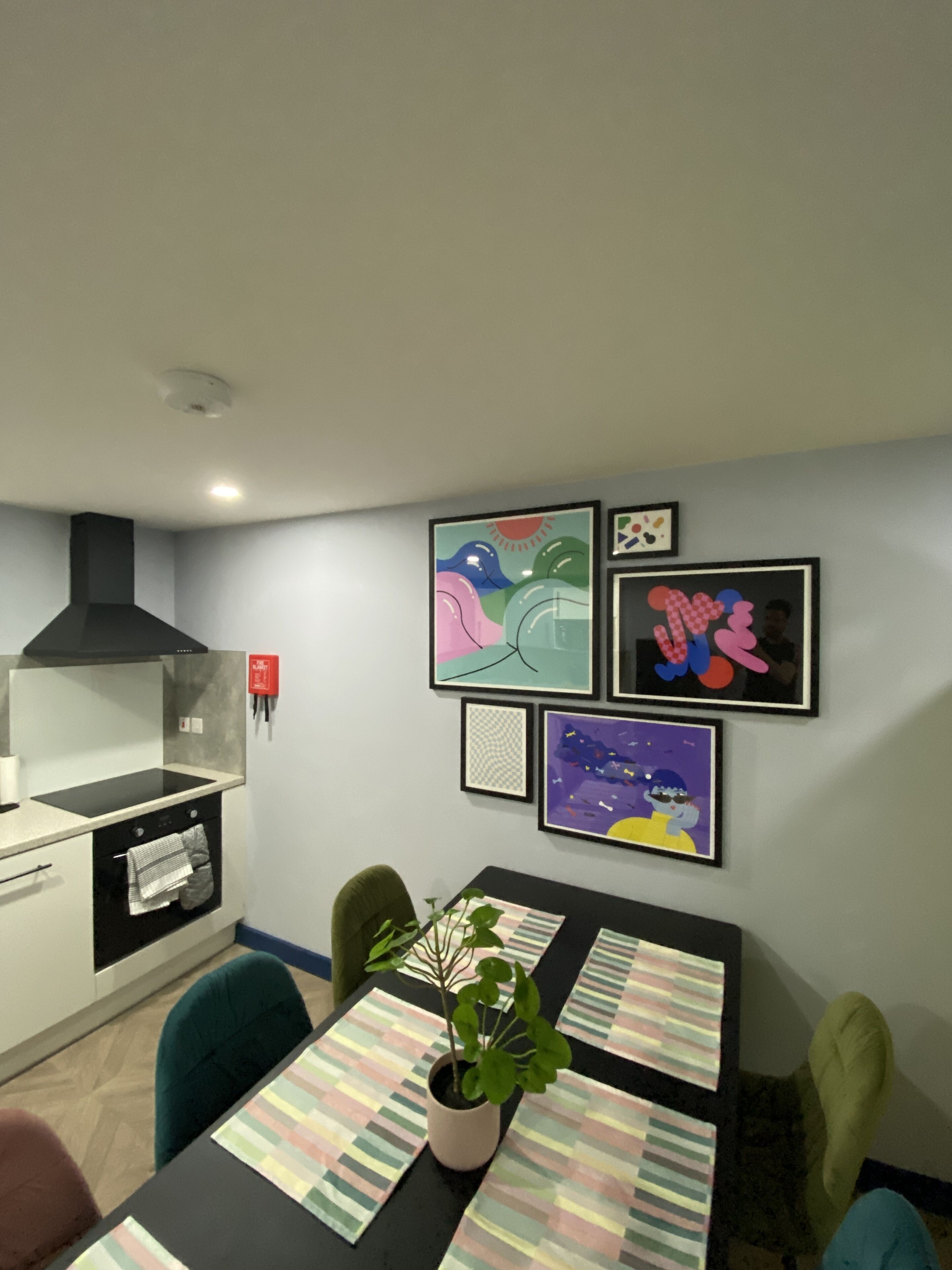 A modern kitchen with a stove, oven, and black range hood. There is a dining table with striped placemats and a potted plant. The walls are adorned with colorful framed artwork.