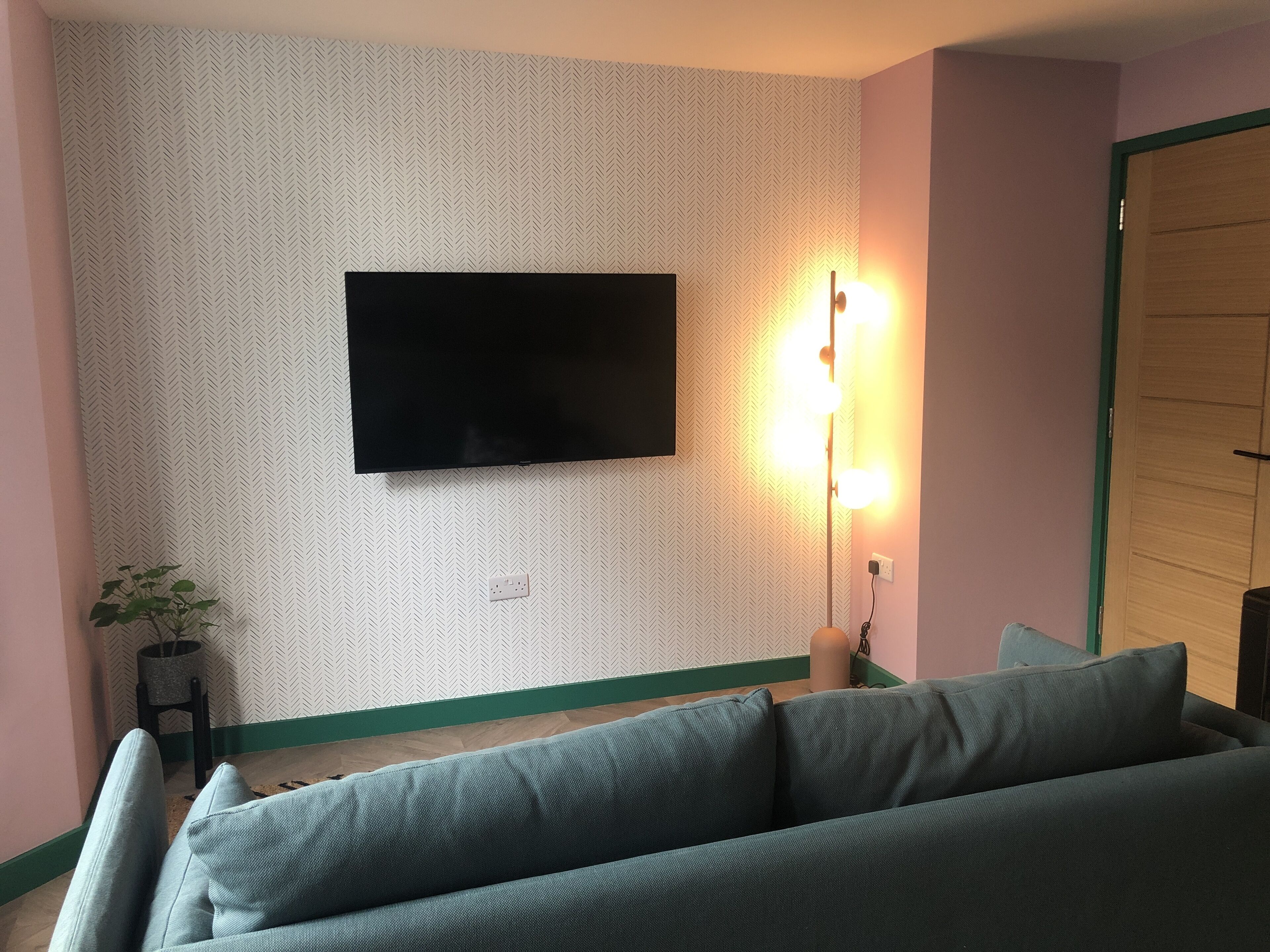 A room with a flat-screen TV mounted on a patterned wall, a green-framed sofa, and a small table with a plant.
