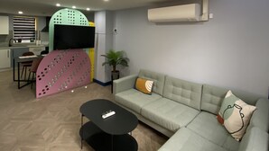 En moderne stue med en hjørnesofa, et sort sofabord og et fjernsyn. I baggrunden er der et køkkenområde med et spisebord og stole.