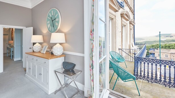 A room with a wooden desk, two lamps, a clock, and a view of a balcony with chairs. - The Zetland Apartment (Saltburn-by-the-Sea)